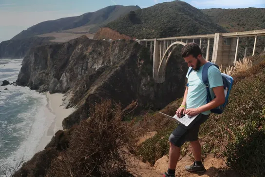 A man working on a laptop, standing on a cliff above the ocean in front of a large arch bridge.